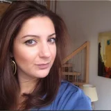 Close-up of woman with long brown hair in blue top, wooden staircase in background.